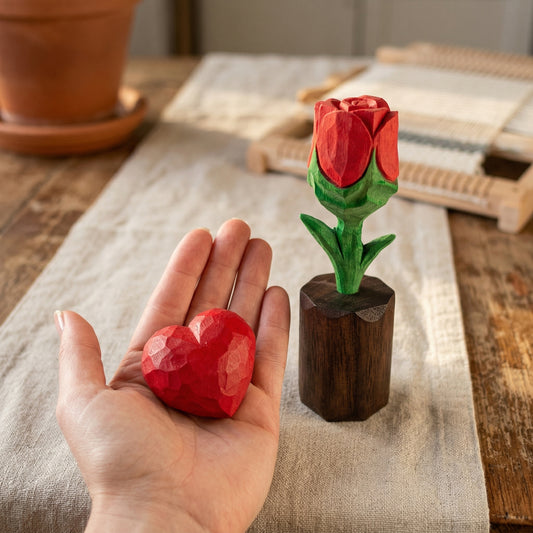 Hand holding a red heart-shaped object next to a wooden rose on a textured surface.