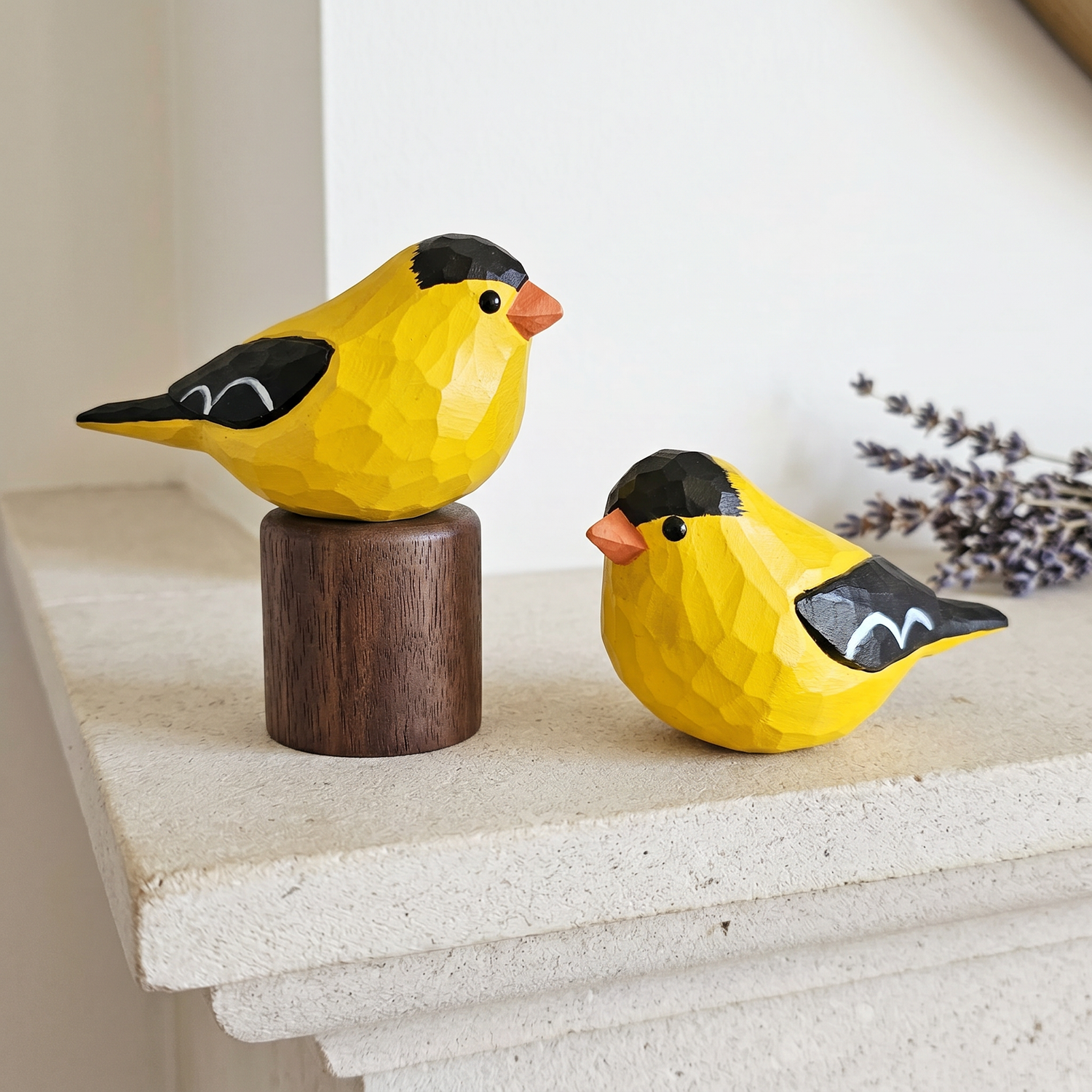 Two hand-painted American Goldfinch wooden figurines on a stone surface decorated with dried lavender sprigs