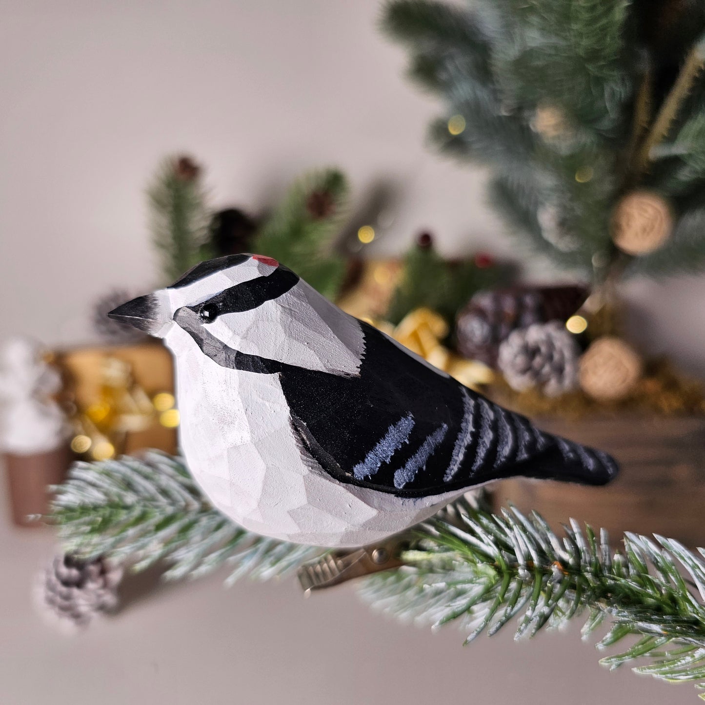 Hand-carved solid basswood Downy Woodpecker clip-on ornament perched on a pine branch, showing black-and-white hand-painted plumage and red crown detail