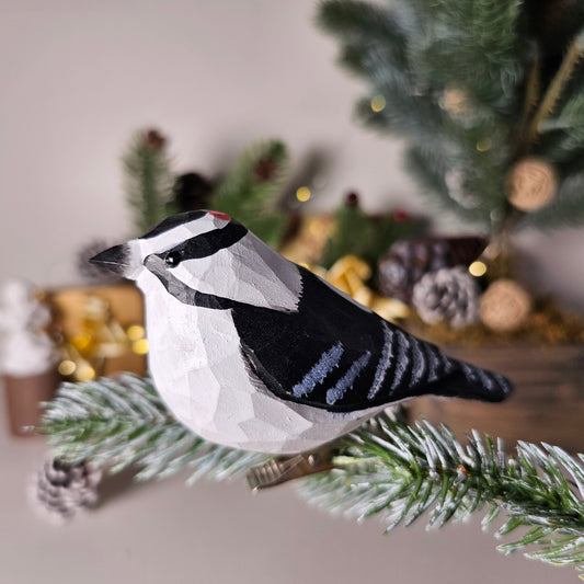Hand-carved solid basswood Downy Woodpecker clip-on ornament perched on a pine branch, showing black-and-white hand-painted plumage and red crown detail