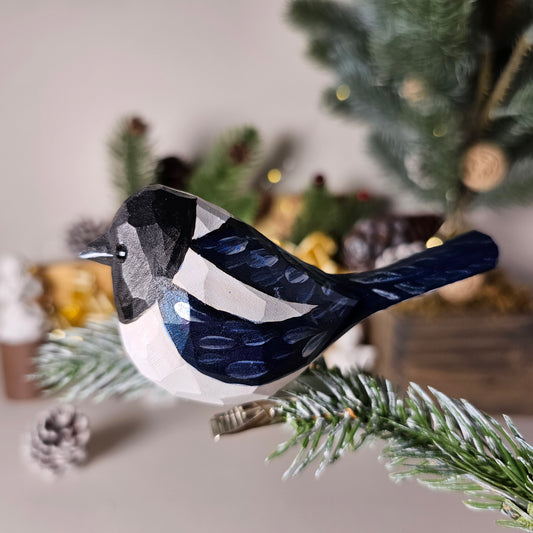 Hand-carved solid basswood Eurasian Magpie clip-on bird ornament, showing bold black-and-white plumage and iridescent tail, clipped to a pine branch