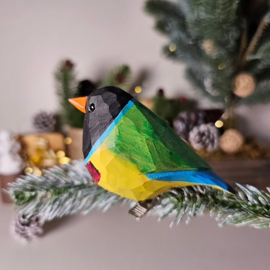 Hand-carved solid basswood Gouldian Finch clip-on ornament showing vivid red head, purple breast, and golden-yellow belly hand-painted detail, perched on a pine branch