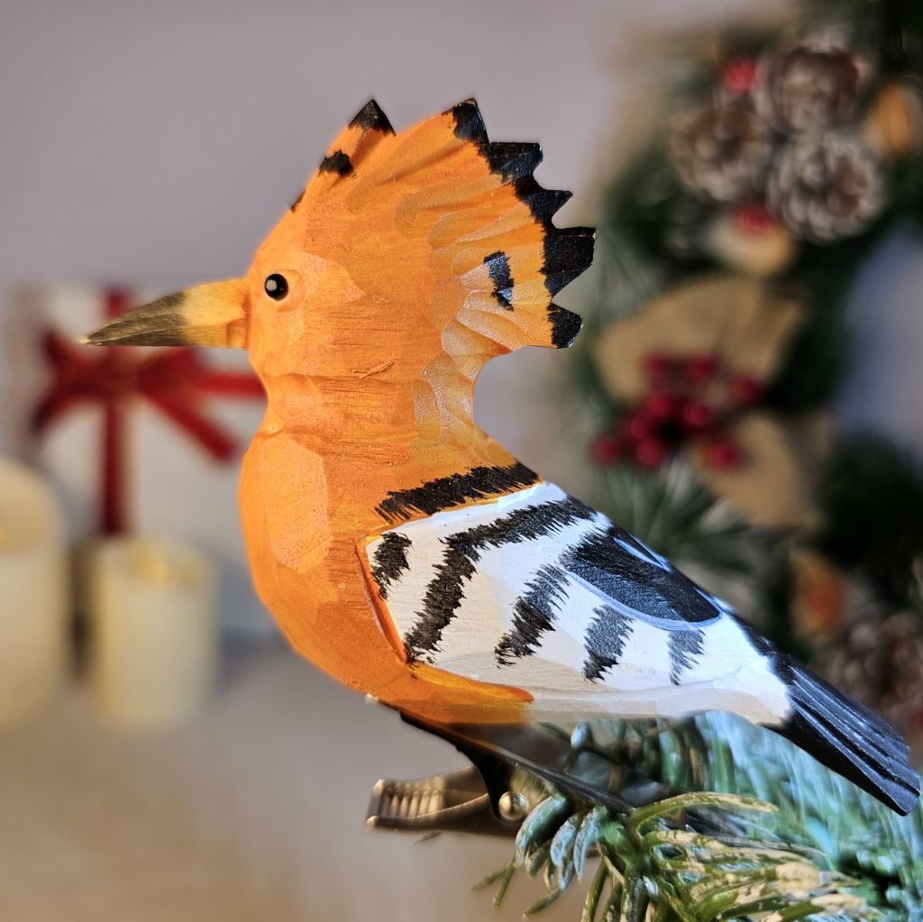 Close-up of the spring clip mechanism on a hand-painted Hoopoe wooden ornament attached to a Christmas tree branch, showing secure mount and feather detail.