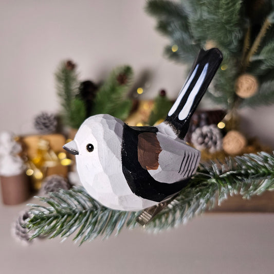 Hand-carved solid basswood Long-tailed Tit clip-on bird ornament, showing blush-pink and white plumage with elongated tail, clipped to a pine branch