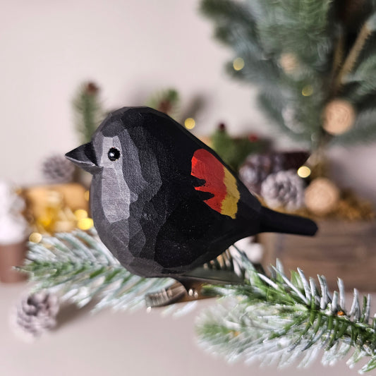 Hand-carved solid basswood Red-winged Blackbird clip-on bird ornament, showing glossy black plumage and vivid scarlet-and-yellow shoulder epaulettes, clipped to a pine branch