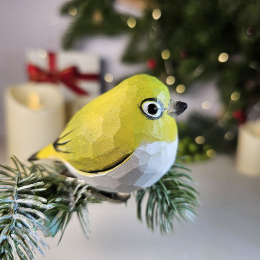 Hand-carved solid basswood Warbling White-eye clip-on ornament showing olive-green plumage and white eye-ring detail, displayed on a pine branch