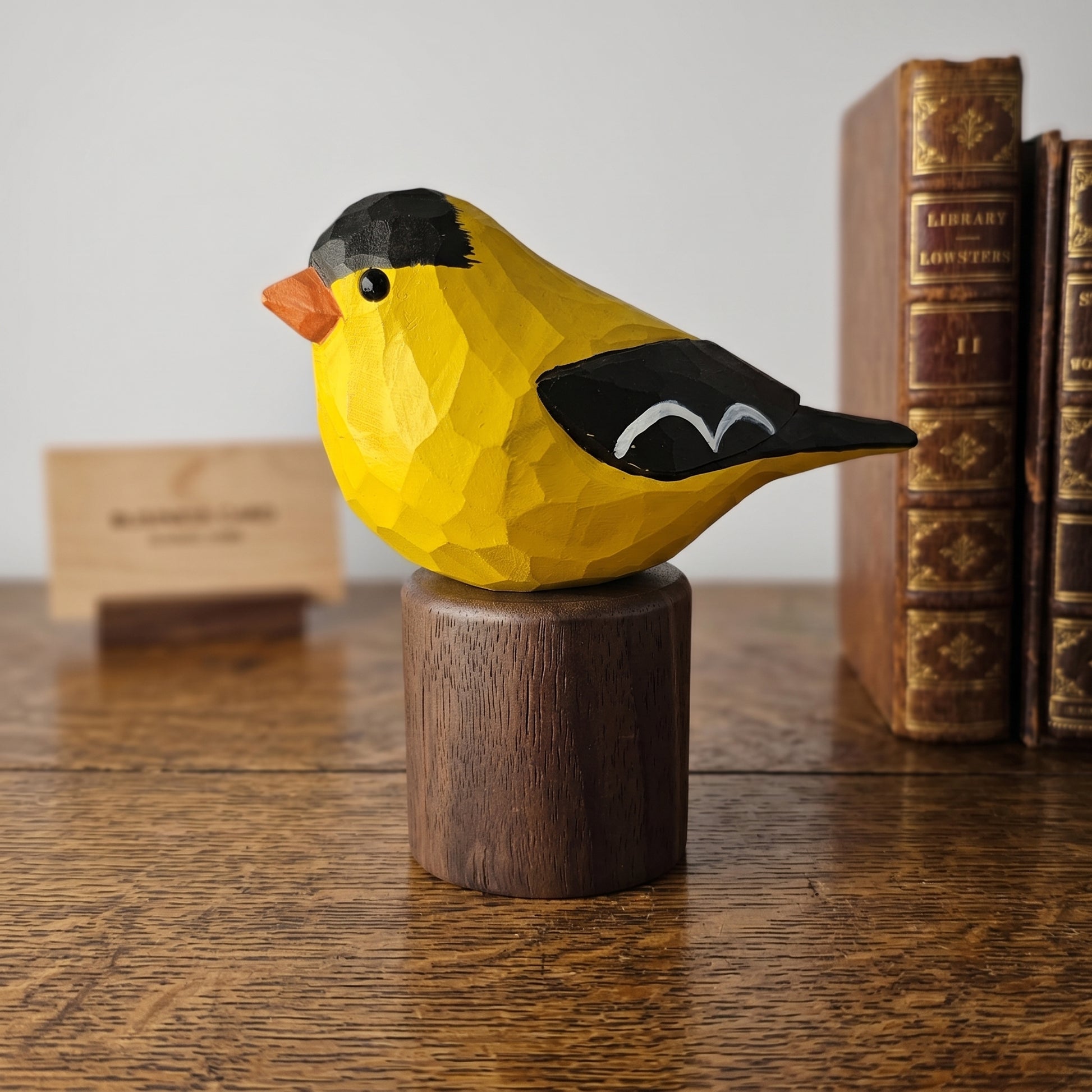 Hand-painted American Goldfinch wood figurine on a dark walnut base, set on a wooden desk next to vintage library books.
