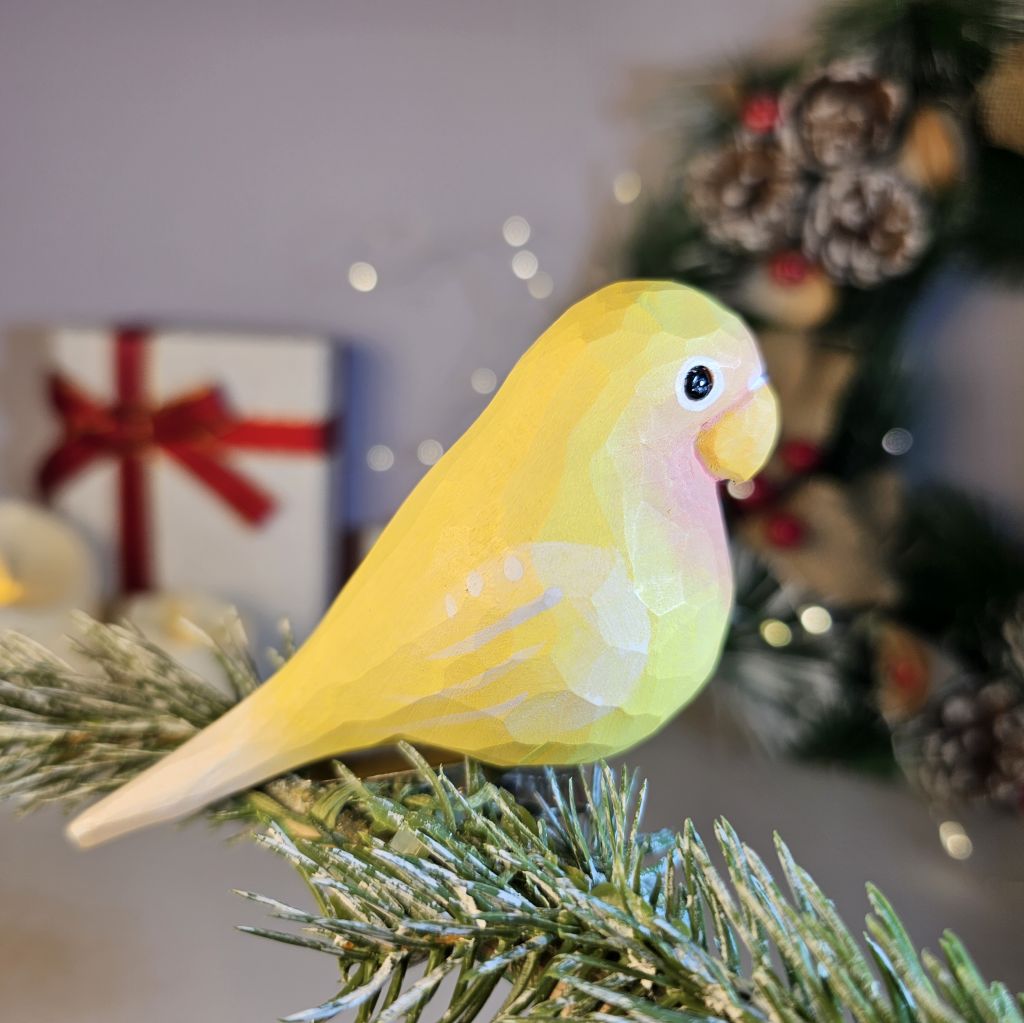 A wooden clip-on Christmas tree ornament painted to resemble a yellow parakeet with a hand-painted detail, attached to a festive holiday decoration backdrop.